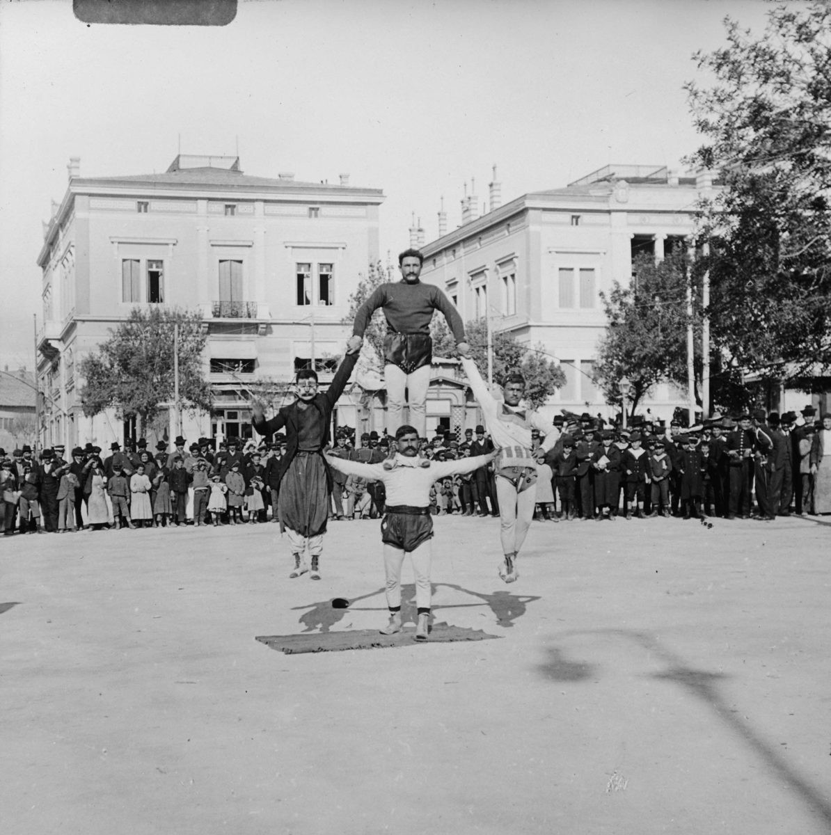 Ακροβάτες στην πλατεία Συντάγματος. Στο βάθος διακρίνονται τα μέγαρα Ιωάννου Βούρου και Στεφάνου Σκουλούδη. Φωτογραφία του Alexander Lamont Henderson.
