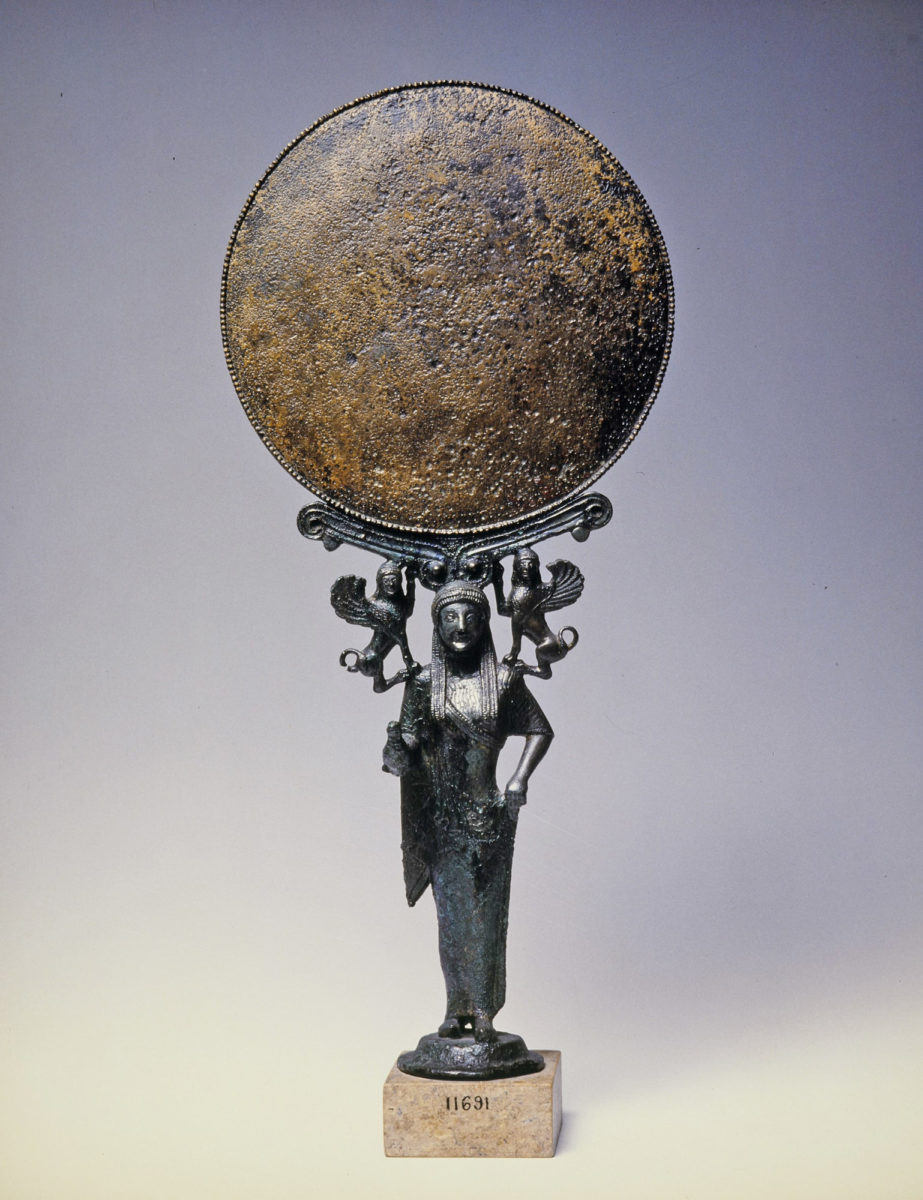 Kάτοπτρο που στηρίζεται σε γυναικεία μορφή, ίσως Aφροδίτη. Φορεί χιτώνα και ιμάτιο και κρατεί πτηνό στο δεξιό χέρι. Στους ώμους της κάθονται δύο σφίγγες. Kορινθιακό εργαστήριο. Γύρω στο 520 π.X. (© Εθνικό Αρχαιολογικό Μουσείο/ TAΠΑ).