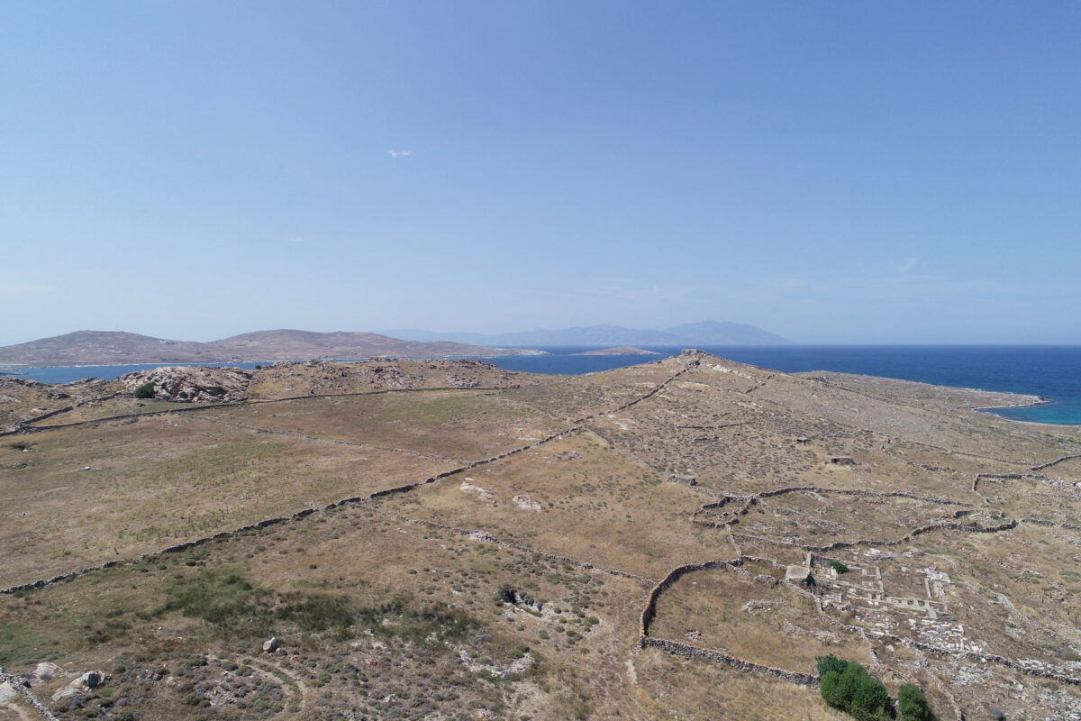 Η ελληνιστική νεκρόπολη της Δήλου στη Ρήνεια, ο πυραμιδοειδής λόφος Χωμασοβούνι όπου πιθανολογείται η θέση του Αρτεμισίου εν νήσω των δηλιακών επιγραφών, και αριστερά αρχαία αγροικία δίπλα σε νεότερη. Στο βάθος, το βόρειο τμήμα της Ρήνειας και στον ορίζοντα η Τήνος (φωτ.: ΥΠΠΟΑ).