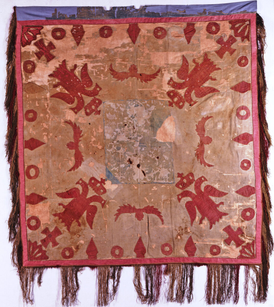 Σημαία Ηπειρωτών που υψώθηκε στον Αγώνα του 1821. Βαμβακερό υφαντό (ΕΙΜ 2744). Φέρει σύμβολα της Βυζαντινής Αυτοκρατορίας: πορφυροί εστεμμένοι αετοί, δικέφαλοι, σταυροί. Στο κέντρο, φθαρμένος πια, δεσπόζει γαλάζιος σταυρός σε λευκό πλαίσιο. © ΙΕΕΕ-ΕΙΜ.