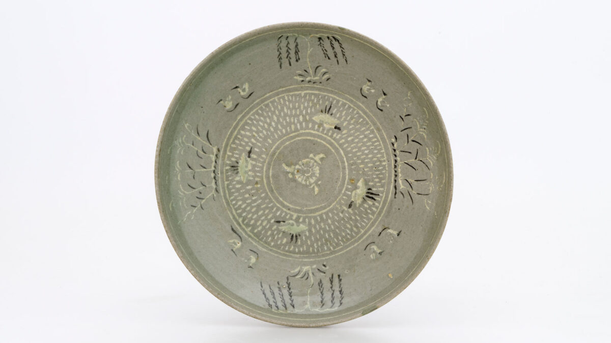Πιάτο. Λιθοκέραμος με διακόσμηση σε λευκό και μαύρο ένθετο αλοίφωμα κάτω από πράσινη εφυάλωση: στο εσωτερικό, ένα άνθος χρυσάνθεμου πλαισιώνεται από στικτό κάμπο γύρω από τέσσερις γερανούς σε πτήση˙ εντός του χείλους, δύο ιτιές και δύο συστάδες ινδοκάλαμων εναλλάσσονται με τέσσερα ζεύγη παπιών ή χηνών που κολυμπούν εντός ταινίας˙ στο εξωτερικό του πιάτου, μια ταινία στικτών κύκλων και μια ταινία με «μαίανδρο» πλαισιώνουν τέσσερα άνθη χρυσάνθεμου γύρω από το πόδι. Κορέα. Δυναστεία Κόρυο (918-1392), όψιμος 13ος-14ος αι. Διάμ. 18,3 εκ. Μουσείο Μπενάκη 2417. Δωρεά Γεωργίου Ευμορφόπουλου.