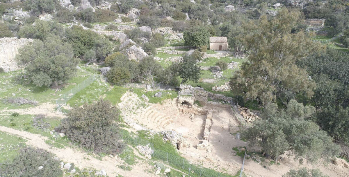 Άποψη του αρχαίου ωδείου από τα νότια του αρχαιολογικού χώρου της Λισού (στο βάθος ο βυζαντινός ναός Άι-Κυρκού). Φωτ.: ΥΠΠΟΑ.