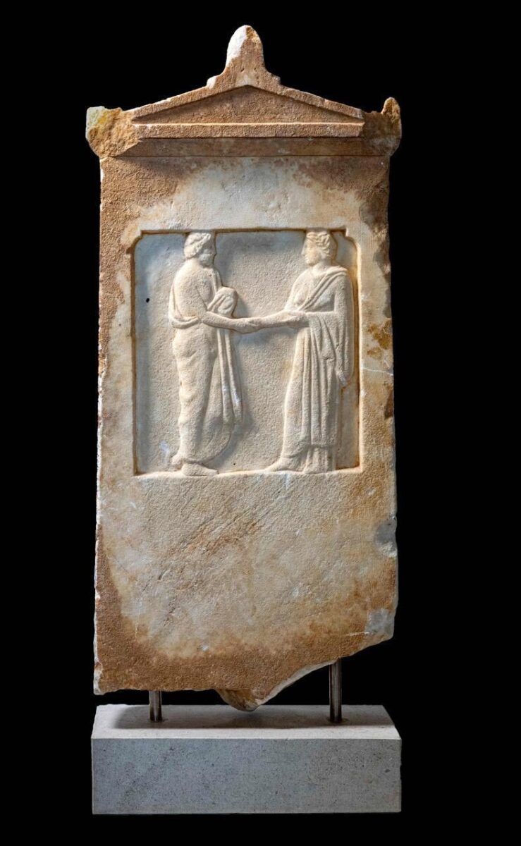 Μαρμάρινη αττική επιτύμβια στήλη με αετωματική επίστεψη. Φέρει ανάγλυφη παράσταση σε ορθογώνιο διάχωρο. Απεικονίζεται σκηνή δεξίωσης ανάμεσα σε ανδρική και γυναικεία μορφή. Ύψος 90,2 εκ. Αρχές 4ου αι. π.Χ. Πηγή εικόνας: ΥΠΠΟ.