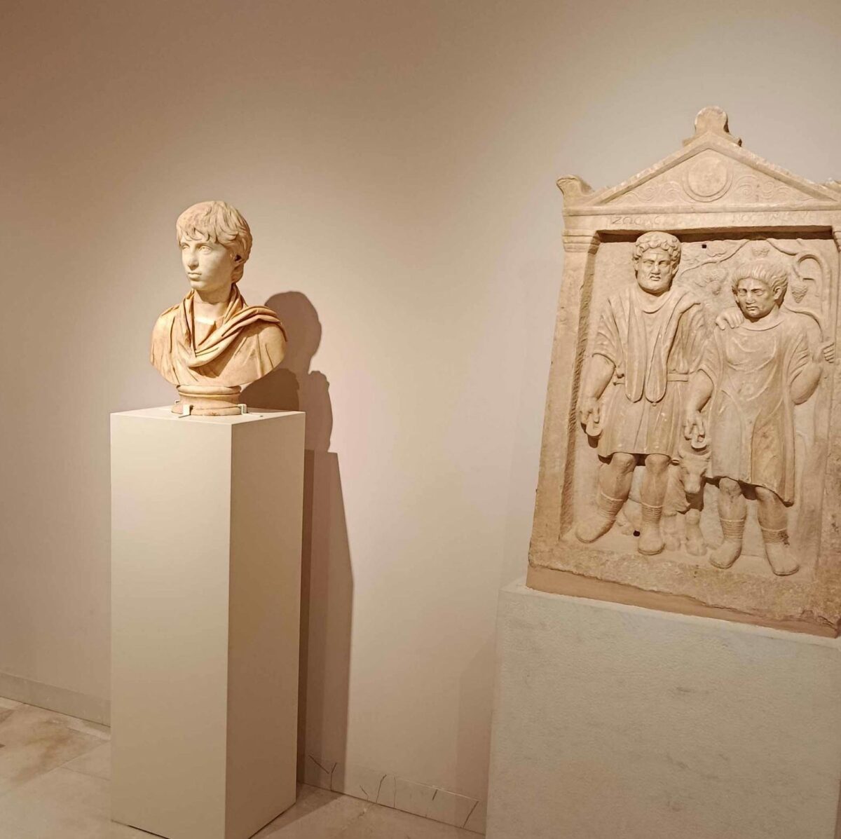 Ένα από τα νέα εκθέματα του Αρχαιολογικού Μουσείου Μαραθώνα. Αριστερά, μαρμάρινη προτομή με πορτρέτο του Πολυδευκίωνα, μαθητή του Ηρώδη Αττικού (2ος αι. μ.Χ.) που βρέθηκε στο Ιερό των Αιγυπτίων Θεών στη Μπρεξίζα. Πηγή εικόνας: Εφορεία Αρχαιοτήτων Ανατολικής Αττικής.